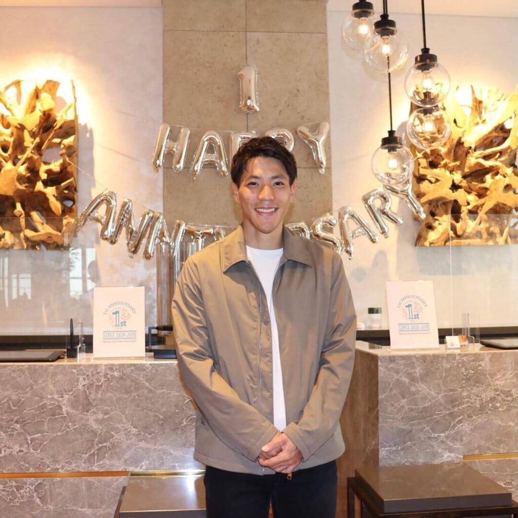誕生日のバルーンの前で笑顔で手を組んでたつカジュアルな服装の山縣亮太